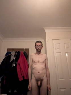 Shirtless man with glasses and short beard standing in a room with coats hanging on hooks and a white door behind him.