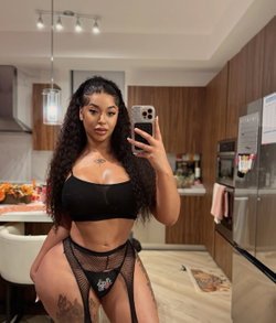 A woman with long curly hair takes a mirror selfie in a kitchen, wearing a black crop top and mesh thong underwear,