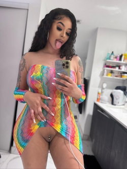 Curvy woman with dark curly hair and tattoos takes a mirror selfie in a colorful mesh dress, sticking out her tongue in