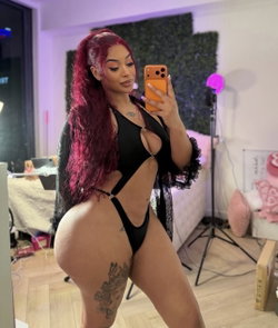 Woman with long red hair in a black cutout bodysuit and lace robe takes a mirror selfie in a room with pink lighting
