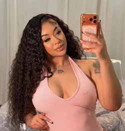 Curvy woman with long dark curls takes a mirror selfie in a pink halter top, showing tattoos on her chest and arm.