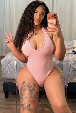 Curvy woman with long dark curls wears a pink one-piece swimsuit, poses in a mirror selfie with visible thigh tattoos,