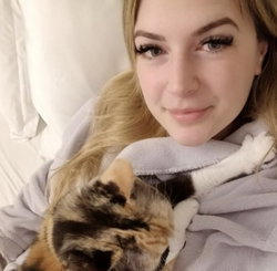 Blonde woman with long hair smiles softly while holding a calico cat, wrapped in a gray blanket on a bed.