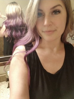 Woman with purple-tipped hair in a black tank top takes a mirror selfie in a bathroom.