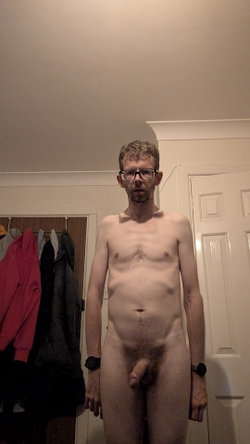 A shirtless man with glasses and a beard stands nude in a room with coats hanging on the wall behind him.