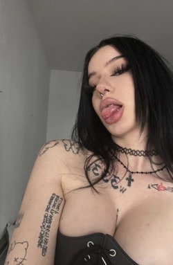 A woman with long black hair, multiple tattoos on her arms and chest, wearing a black corset top and choker, sticking
