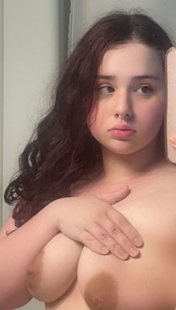 A woman with long dark hair and a nose ring takes a mirror selfie, covering her chest with one hand in a bathroom