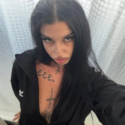 A woman with long black hair and visible chest tattoos takes a selfie in a black zip-up hoodie, standing near a