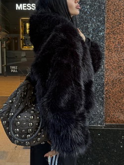 Woman in black faux fur coat and studded bag, standing outdoors near a storefront.