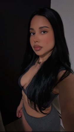 A woman with long black hair and a curvy figure poses in a gray crop top and matching bottoms, standing indoors against