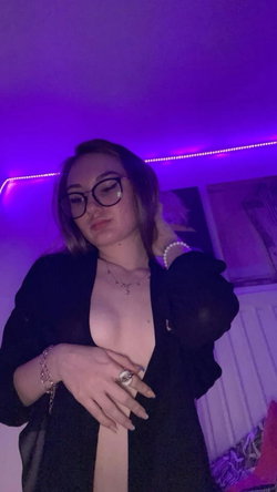 A woman with dark hair and glasses poses in a dimly lit room with purple LED lighting, wearing an open black top that