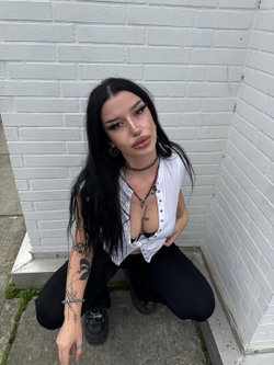 A woman with long black hair and tattoos crouches against a white brick wall, wearing a white unbuttoned top over a bra