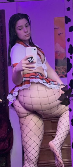 Brunette woman in a plaid skirt and fishnet stockings takes a mirror selfie in a purple-lit room.