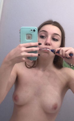 A young woman with long hair and dark nails brushes her teeth in a bathroom selfie.