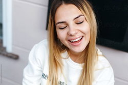 A smiling woman with long blonde hair wearing a white sweatshirt, captured in a close-up selfie-style shot indoors.