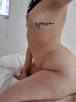 A woman with dark hair and visible tattoos on her side and wrist is sitting on a bed, partially nude, with bare breasts