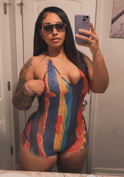 Curvy woman with long dark hair and sunglasses takes a mirror selfie in a colorful one-piece swimsuit, showing tattoos
