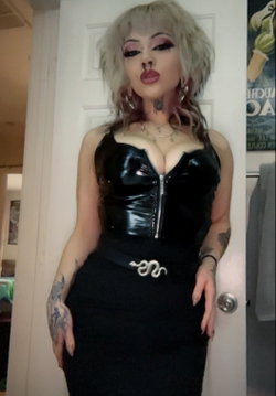 A woman with platinum blonde hair and bold makeup wears a black latex corset top and high-waisted skirt, posing indoors