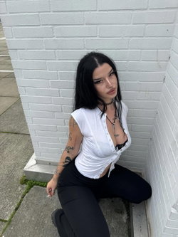 A woman with long black hair and multiple arm tattoos kneels against a white brick wall, wearing a white button-up top