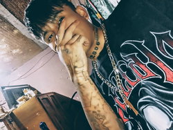 A young man with short dark hair and tattoos on his neck and arm wears a black graphic tee and gold chain, posing