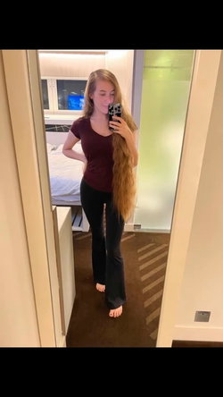 A woman with long blonde hair takes a mirror selfie in a maroon top and black flared pants, standing barefoot in a