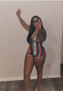 Curvy woman with long dark hair and sunglasses poses in a colorful one-piece swimsuit indoors, arm raised against a