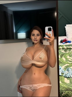 Curvy woman in beige lace lingerie takes a mirror selfie in a hotel room, holding her phone with one hand.