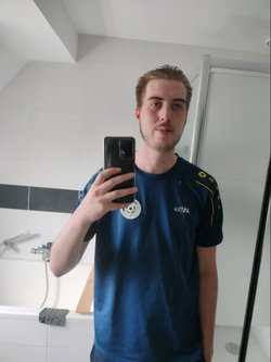 A young man with light hair and a beard takes a mirror selfie in a bathroom, wearing a blue athletic shirt.
