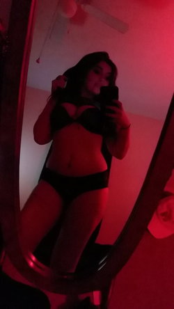 A woman with long dark hair takes a mirror selfie in black lingerie under red lighting, showing a curvy figure.
