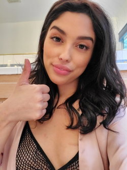 Brunette woman with wavy hair gives a thumbs-up in a bathroom, wearing a black mesh top under a pink robe.