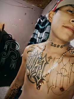 Shirtless person with extensive chest and neck tattoos, wearing a gold chain and cap, posing indoors near a wall with
