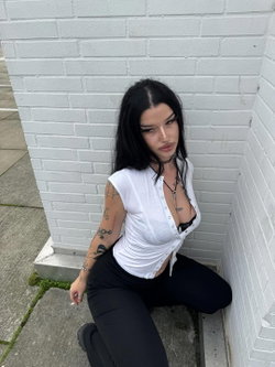 A woman with long black hair and tattoos on her arm kneels against a white brick wall, wearing a white button-up top
