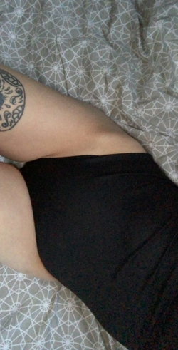 Close-up of a person's arm with a circular tattoo resting on a patterned bedsheet, wearing black fabric.