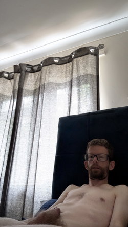 A shirtless man with glasses and a beard lies on a bed with a dark headboard, in a room with gray curtains and natural
