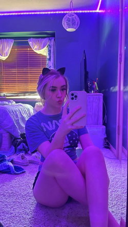 Blonde woman in a graphic tee and shorts sits on the floor, taking a mirror selfie with cat ears under purple LED