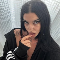 A woman with long dark hair and multiple tattoos on her chest and shoulder wears a black hoodie, posing indoors with a