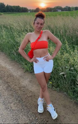 Curvy woman in red strapless top and white shorts poses confidently in a field at sunset, wearing white sneakers.