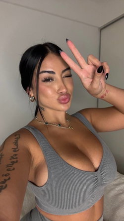 Brunette woman with tattoos poses in a gray ribbed crop top, making a peace sign and puckering her lips in a