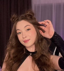 Brunette woman with wavy hair styled in buns, wearing a black choker and long-sleeve top, posing indoors with hand in