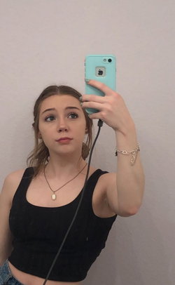 A young woman with light brown hair takes a mirror selfie in a black crop top and jeans, holding a teal phone.