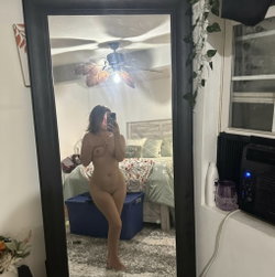 A woman with dark hair stands nude in a bedroom, taking a mirror selfie while covering her chest; the room includes a