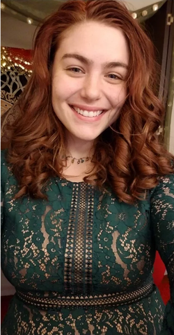 A woman with wavy red hair smiles warmly, wearing a green lace dress indoors with festive lights in the background.