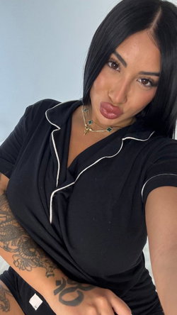 Brunette woman with tattoos in black pajama top, posing closely for a selfie against a plain background.