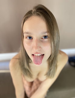 A young woman with light brown hair and blue eyes sticks her tongue out playfully while topless, hands covering her