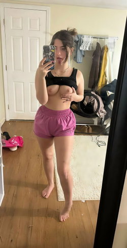 A woman with dark hair takes a mirror selfie in a black crop top and pink shorts, holding her phone with one hand and
