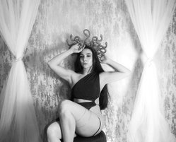 A woman with long dark hair poses in a one-shoulder top and thong, wearing a snake crown, against a textured wall with