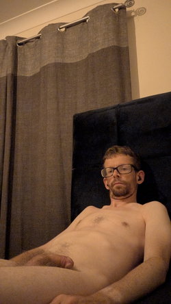 A shirtless man with glasses and a beard lies on a dark headboard, partially nude, in a dimly lit room with gray