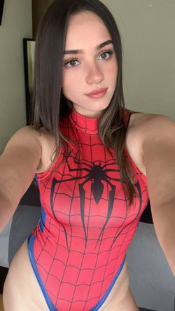 Brunette woman in a red and blue Spider-Man bodysuit poses for a selfie indoors, showing a curvy figure with long hair