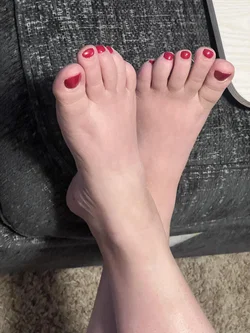 Close-up of bare feet with red toenail polish resting on a dark textured surface, likely a couch or chair.
