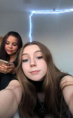Two young women in a room with blue LED lights; one takes a selfie while the other looks at her phone, both wearing